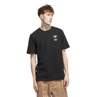 A young man with light brown hair wears a black JY3955 Skate Tee by Adidas Skateboarding, featuring two small trefoil logos on the left chest. He pairs it with beige pants and stands against a plain white background, looking left. Imported product.