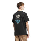 A person stands facing away, wearing a JY3955 Skate Tee Black from Adidas Skateboarding, featuring three stacked trefoil logos in white, gray, and blue on the back; paired with light brown pants.