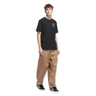 A young man with short, tousled hair wears the black JY3955 Skate Tee from Adidas Skateboarding, paired with tan wide-leg pants and black sneakers with white stripes, standing against a plain white background for an effortlessly cool vibe.