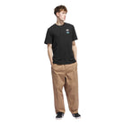 A young man wears the JY3955 Skate Tee Black from Adidas Skateboarding, featuring a small blue and white logo on the left chest, paired with loose tan pants and black sneakers. He has short brown hair and his hands are in his pockets.