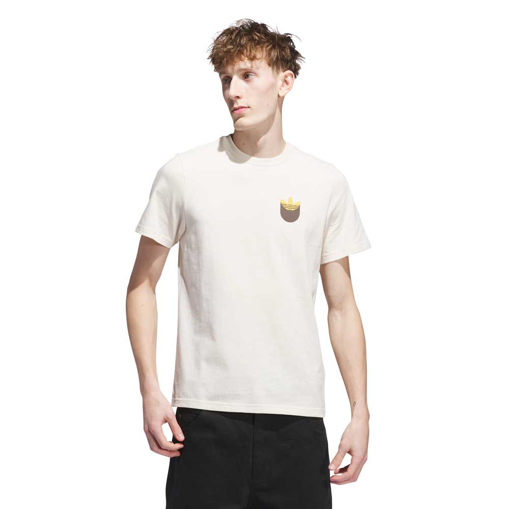 A young man with short, messy hair wears the JY3958 Skate Tee in Wonder White Rustic Orange from Adidas Skateboarding, featuring a small crowned cat graphic on the chest and black pants, standing against a crisp white background.