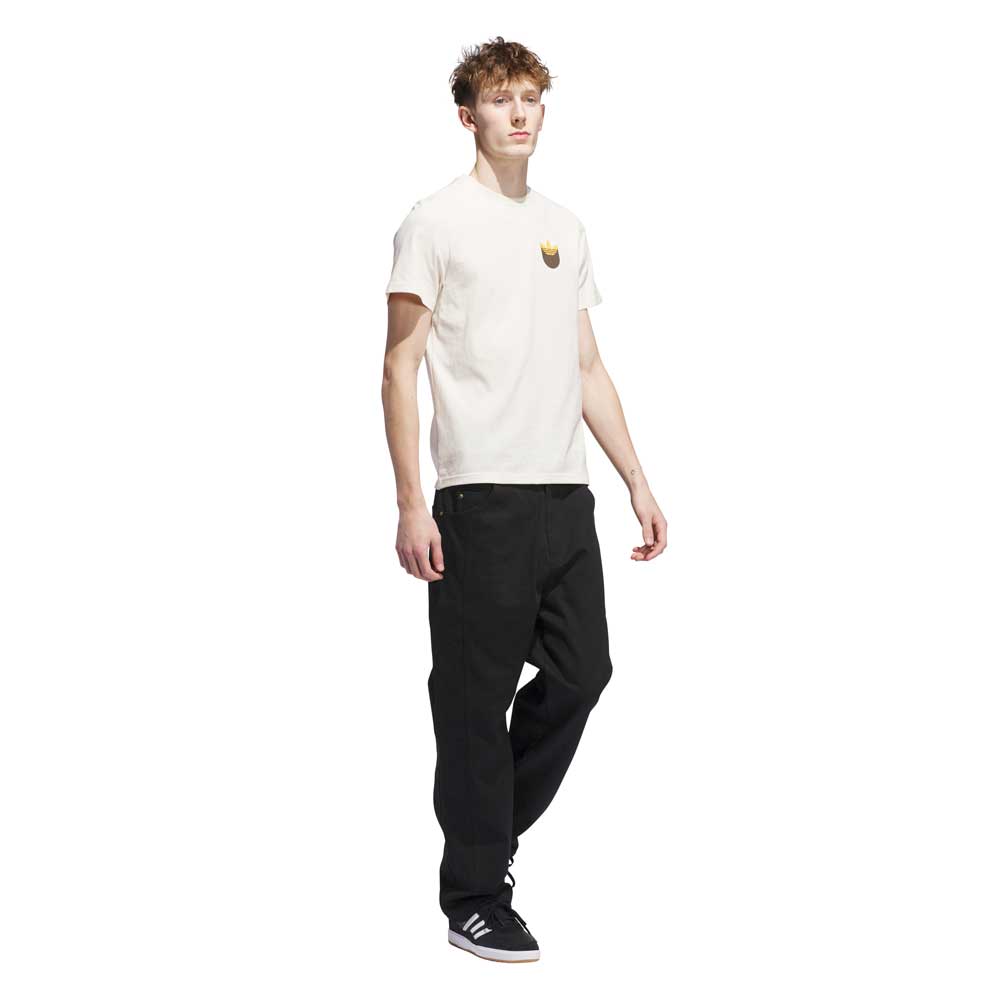 A young man with short, curly hair wears the Adidas Skateboarding JY3958 Skate Tee in Wonder White Rustic Orange, paired with loose black pants and black sneakers, standing against a plain white background.