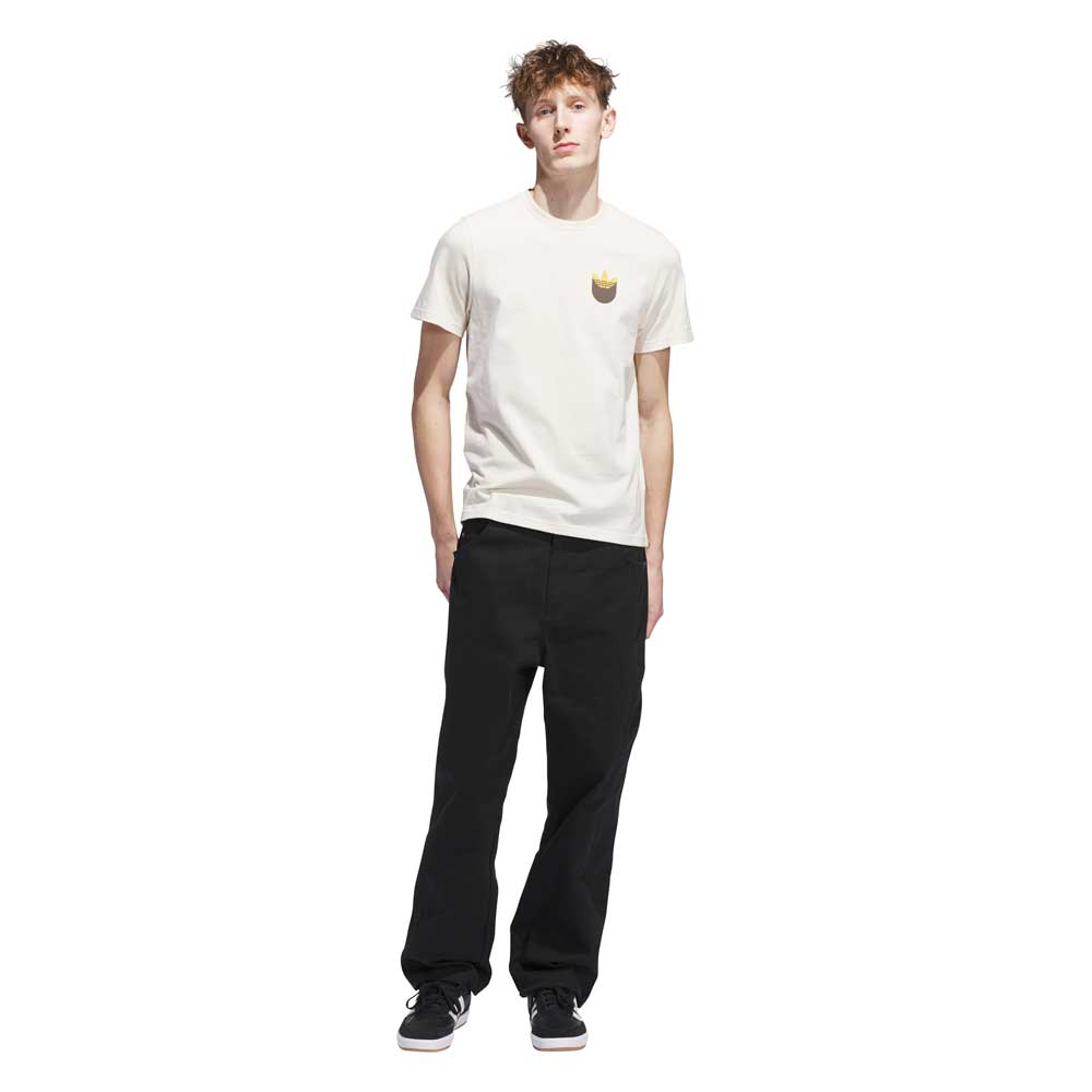 A young man with short, curly hair wears the JY3958 Skate Tee in Wonder White Rustic Orange from Adidas Skateboarding, paired with black pants and sneakers, standing against a plain white background.