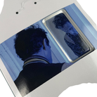 A person with short dark hair and a striped collar is seen from behind, looking into a mirror. Reflected next to them is the Jim Greco Hammers 3 Board Deck Set Collectible, in an image with a cool blue tint.