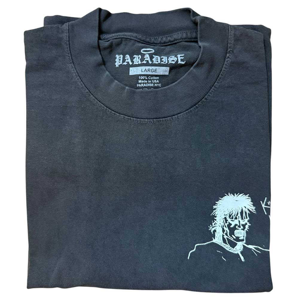 The Kill Kill Kill Vintage Black Tee Paradise is a folded black USA cotton t-shirt featuring a small white line drawing of a spiky-haired character on the lower right, with a PARADISE tag showing size and material details.