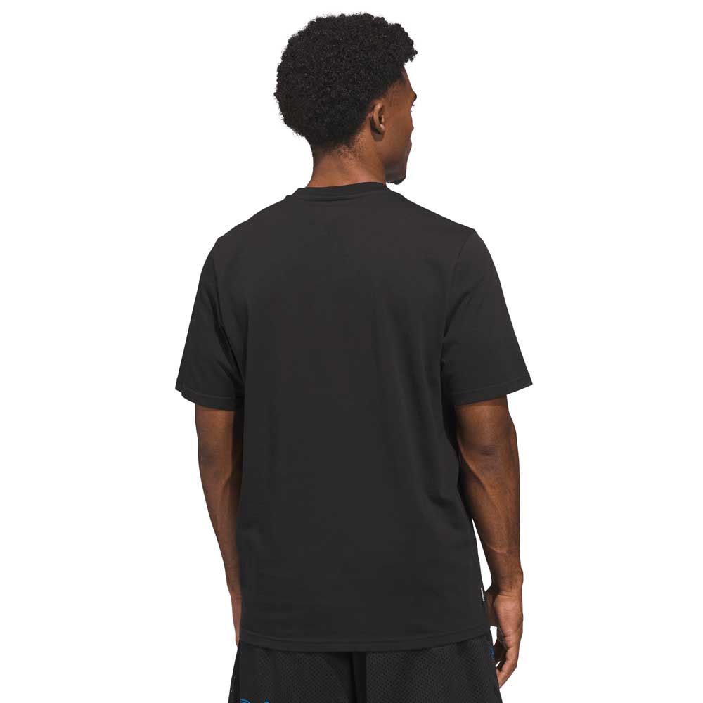 A man with short curly hair stands with his back to the camera, wearing a Kogan Dog Tee Black Blue KC6304 Adidas Skateboarding and black shorts against a white background.