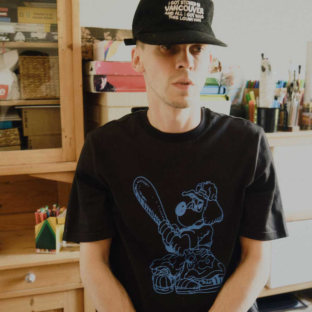 A person wears the Kogan Dog Tee Black Blue KC6304 Adidas Skateboarding and a black cap, standing indoors before shelves filled with books, boxes, and art supplies.