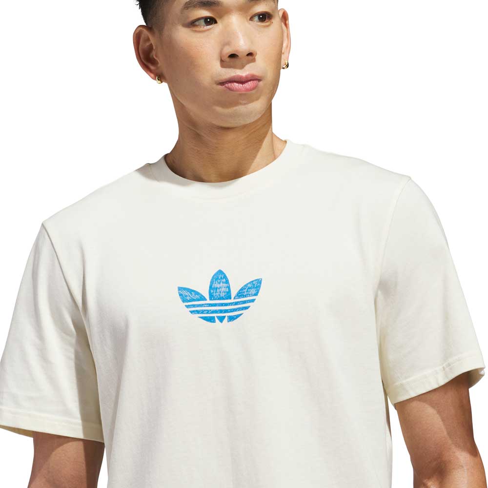 A person wears the Kogan Dragon Tee Cream Blue KC6305 by Adidas Skateboarding, featuring a blue trefoil logo on a cream shirt, looking to the side against a plain white background.