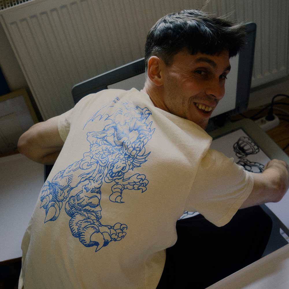 A smiling person glances over their shoulder while drawing at a desk, wearing the Kogan Dragon Tee Cream Blue KC6305 by Adidas Skateboarding, which features an intricate blue dragon graphic on the back.
