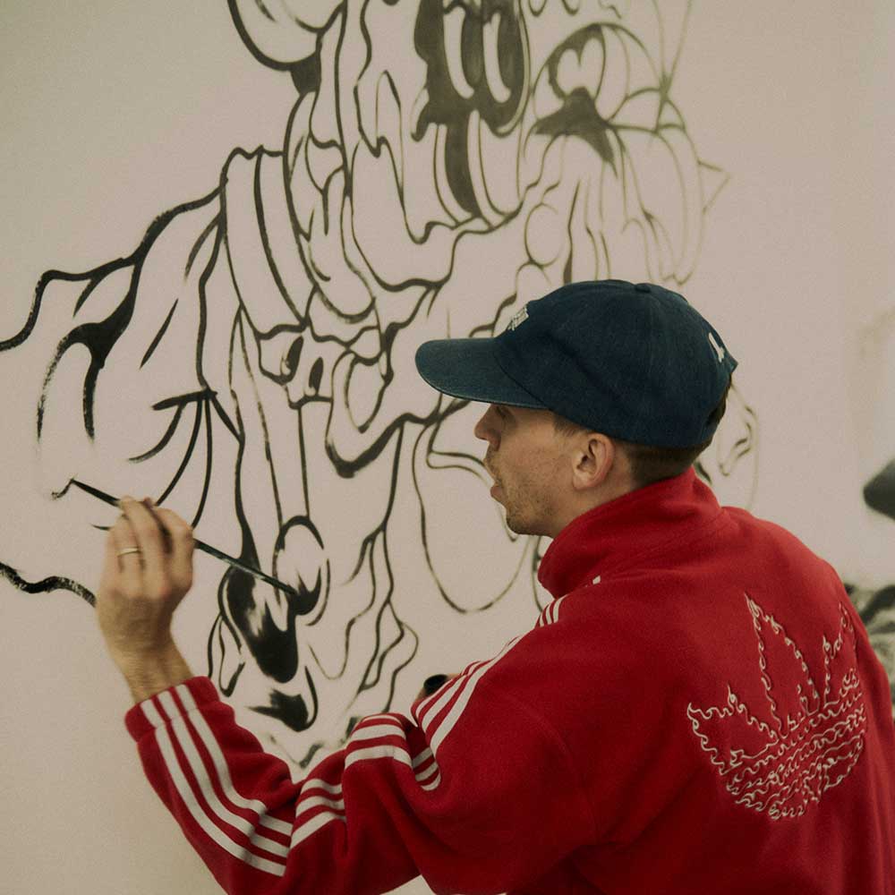 Wearing a Kogan Logo Tee Blue Black KC6301 Adidas Skateboarding, a man in a blue cap draws an abstract black line illustration on a white wall, embodying the creative energy of skate culture.