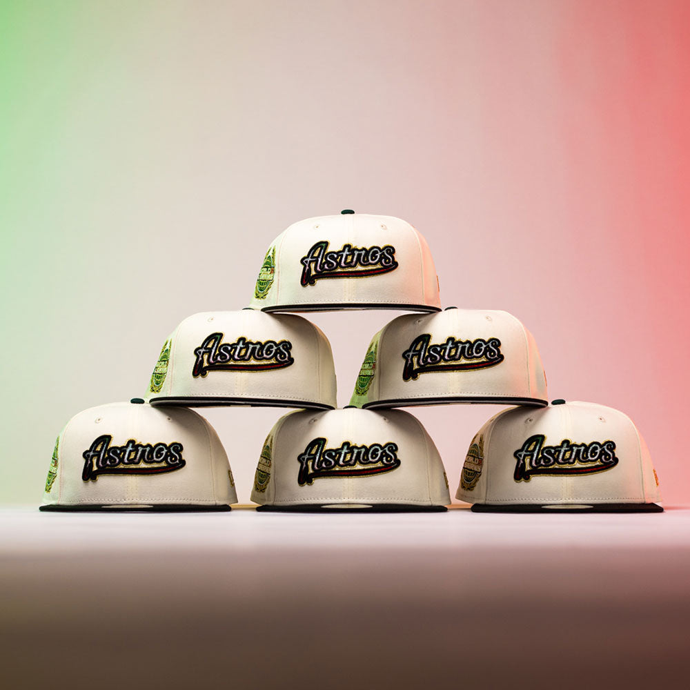 Six La Cumbia WS05 Chrome Black Mexico Houston Astros New Era 5950 Fitted hats, featuring a cream colorway and bold Astros branding—ideal for Houston Astros fans—are stacked in a pyramid arrangement against a backdrop illuminated by gradient red and green lights, creating a dramatic, Chrome Black-inspired display.