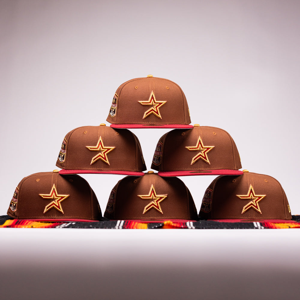 Six Bendicion de la Abuela 20th Brown Red Houston Astros New Era Hat 5950 Fitted caps, featuring yellow star logos, are stacked in a pyramid on a plain background with a colorful cloth at the base.