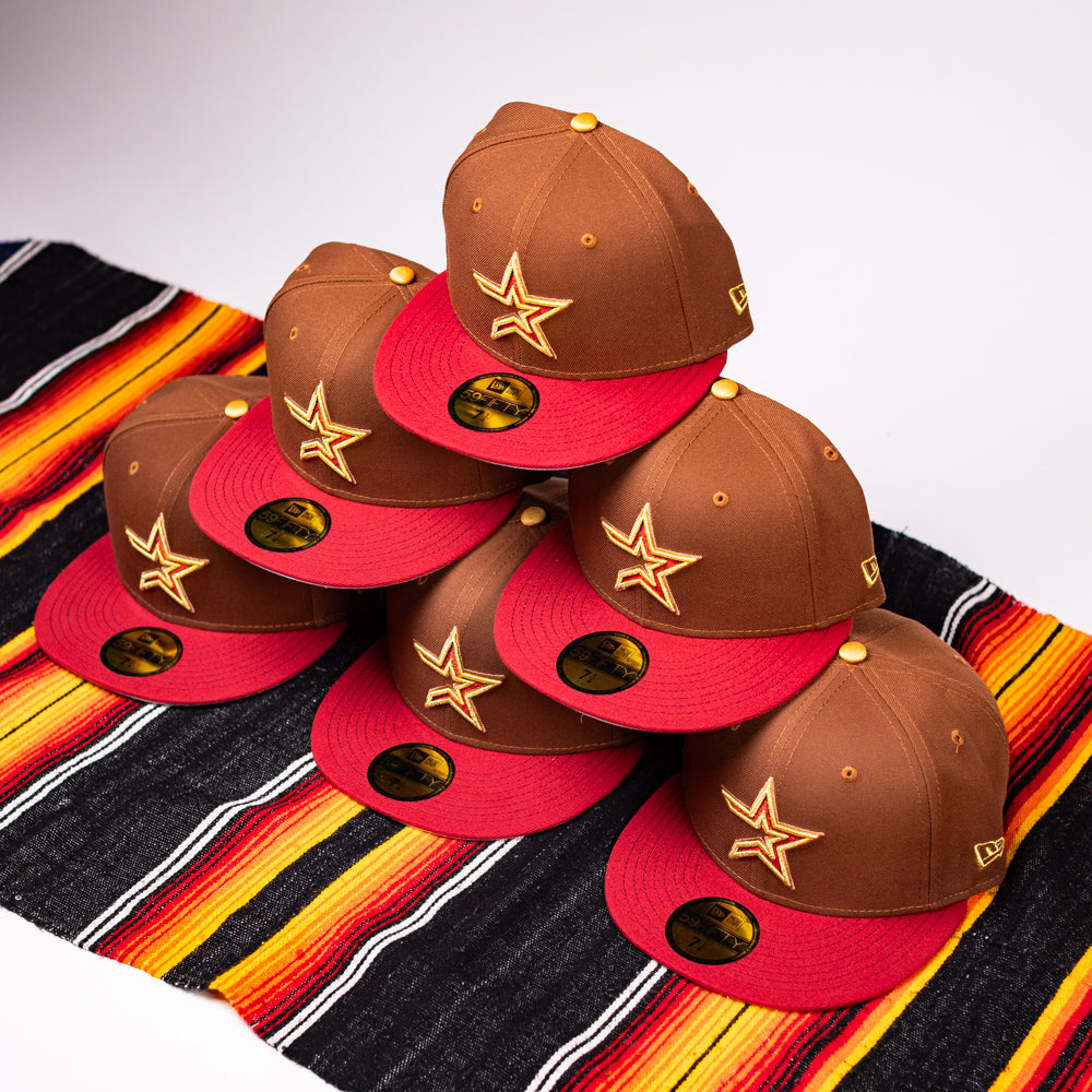 Six Bendicion de la Abuela 20th Brown Red Houston Astros New Era Hat 5950 Fitted caps with yellow star logos are stacked pyramid-style on a colorful striped fabric.