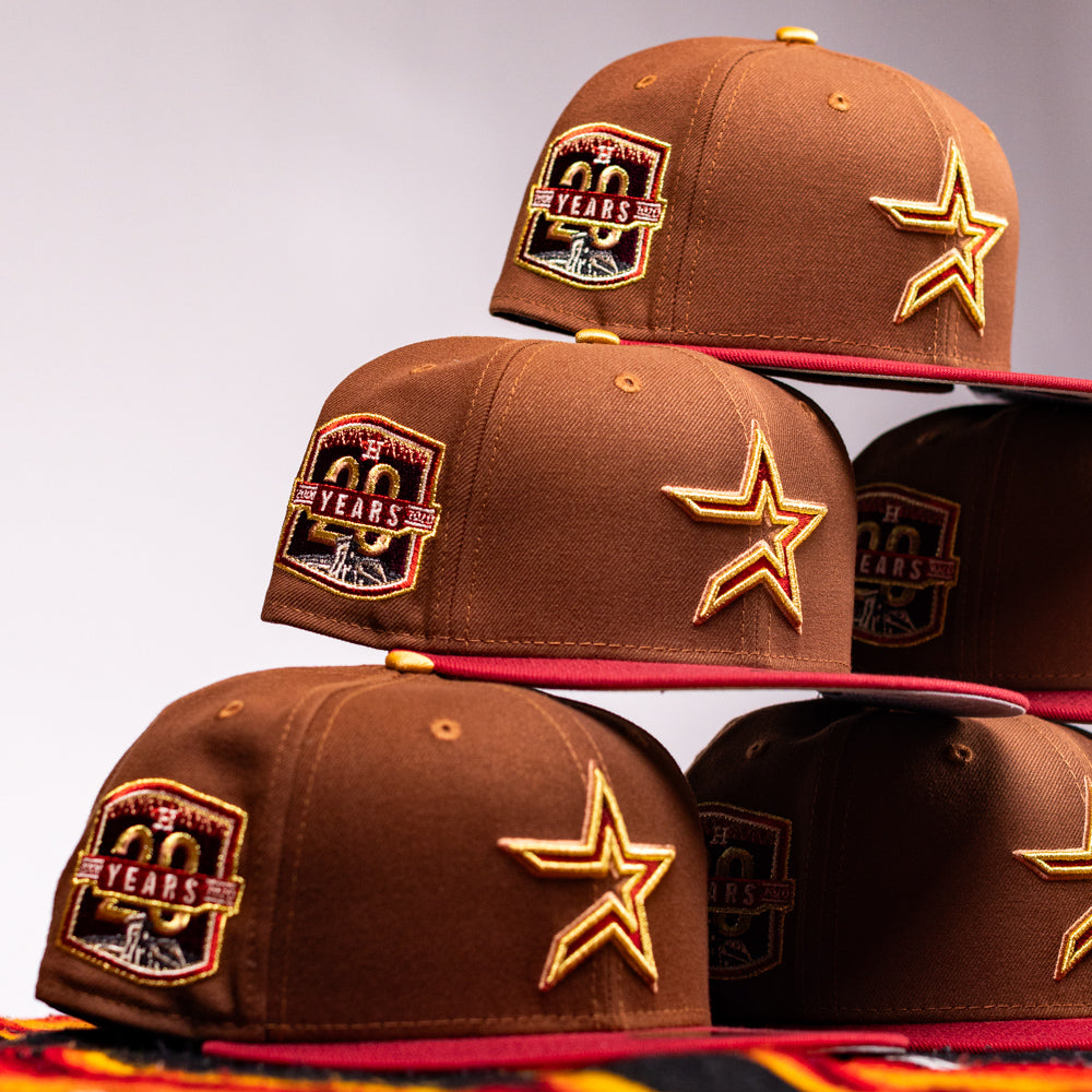 Six Bendicion de la Abuela 20th Brown Red Houston Astros New Era Hat 5950 Fitted hats with gold star and “20 Years” patches are stacked in a pyramid, red brims facing out against a plain light background.