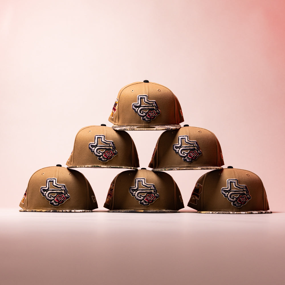Six La Ofrenda Khaki RTC Copa Corpus Christi Hooks New Era 5950 Fitted hats are stacked pyramid-style. Each features an embroidered Texas outline with a rose on the front, set against a softly lit pink and beige background.