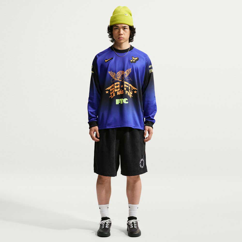 A person stands against a plain white background wearing a lime green beanie, black shorts, white socks, black sneakers, and the Long Sleeve Skate Top IQ6594514 from Nike SB Skateboarding.