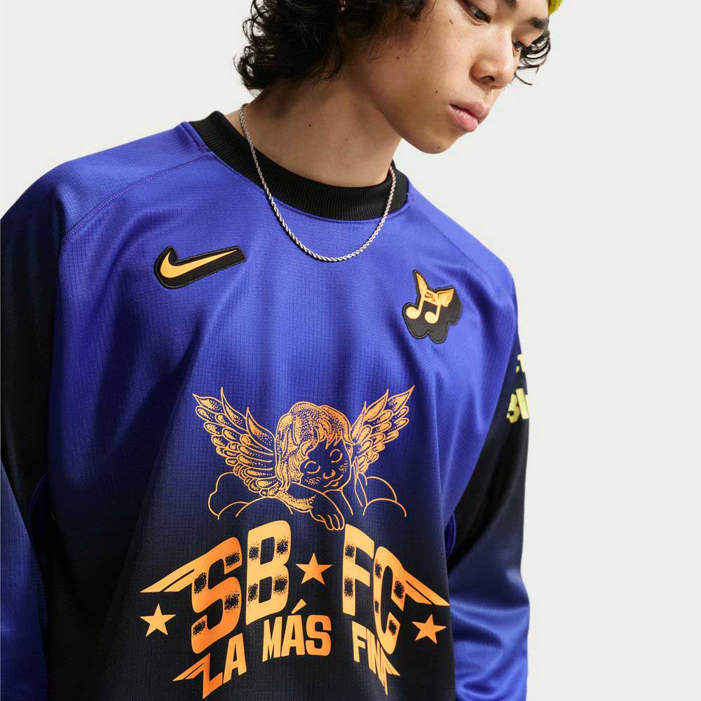 A person is looking down wearing the Nike SB Skateboarding Long Sleeve Skate Top IQ6594514 with blue, black, and yellow graphics including an angel, stars, musical notes, and "SB LA MÁS FINA FC" text. Features Nike and other logos.