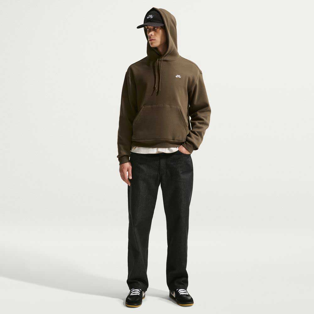 A person in a brown 100% cotton hoodie, Loose Denim Black Pant IB2992010 Nike SB, black Nike SB sneakers, and a black cap stands against a plain white background, looking to the side with one hand in their pocket.