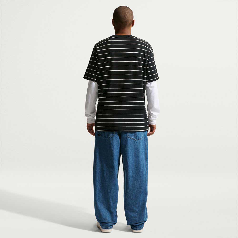 A person stands facing away from the camera, wearing a black and white striped tee over a long-sleeve white shirt, Loose Denim French Blue Pant II7363487 Nike SB jeans, and Nike SB sneakers against a plain white background.