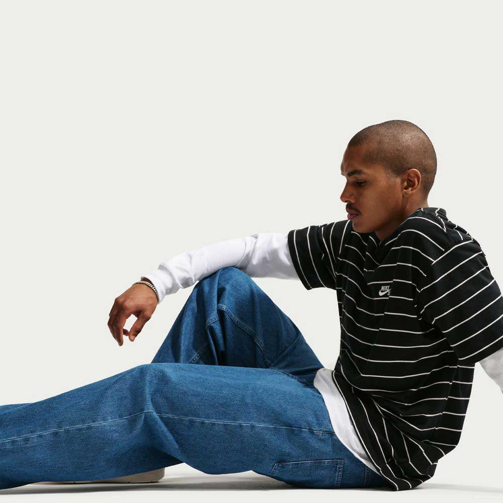 A person with a shaved head sits on the floor against a plain background, wearing a black-and-white striped shirt over a white long-sleeve and Loose Denim French Blue Pant II7363487 Nike SB. They appear relaxed and thoughtful.