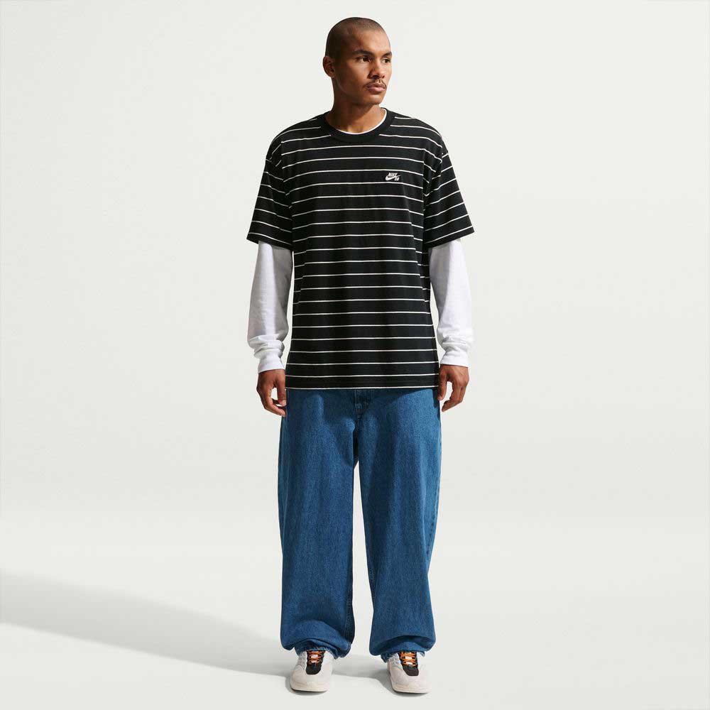 A person stands slightly turned, wearing a black and white striped 100% cotton tee over a long-sleeved white shirt, Loose Denim French Blue Pant II7363487 by Nike SB, and Nike SB white sneakers against a plain light background.