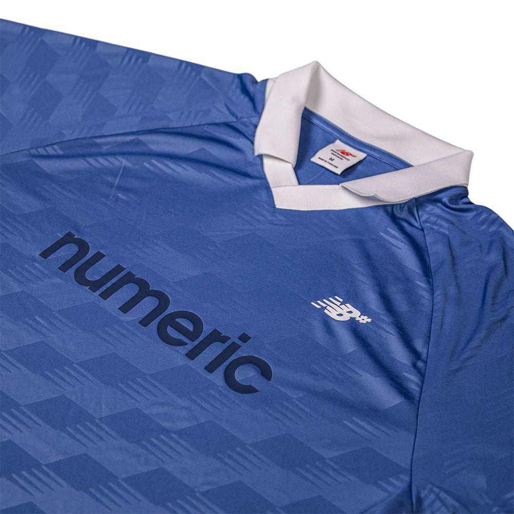 A close-up of the New Balance Numeric Team Jersey MT61V471ABR: a blue soccer jersey with a white collar, “numeric” in dark blue text, the New Balance logo on the right chest, and a subtle geometric fabric pattern.