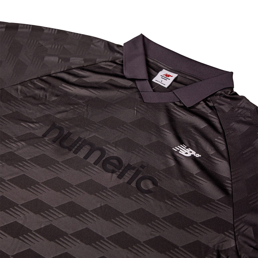 A close-up of the New Balance Numeric Team Jersey MT61V471ABR in dark gray, featuring a subtle geometric pattern, a numeric logo across the chest, and the New Balance logo on the right side.