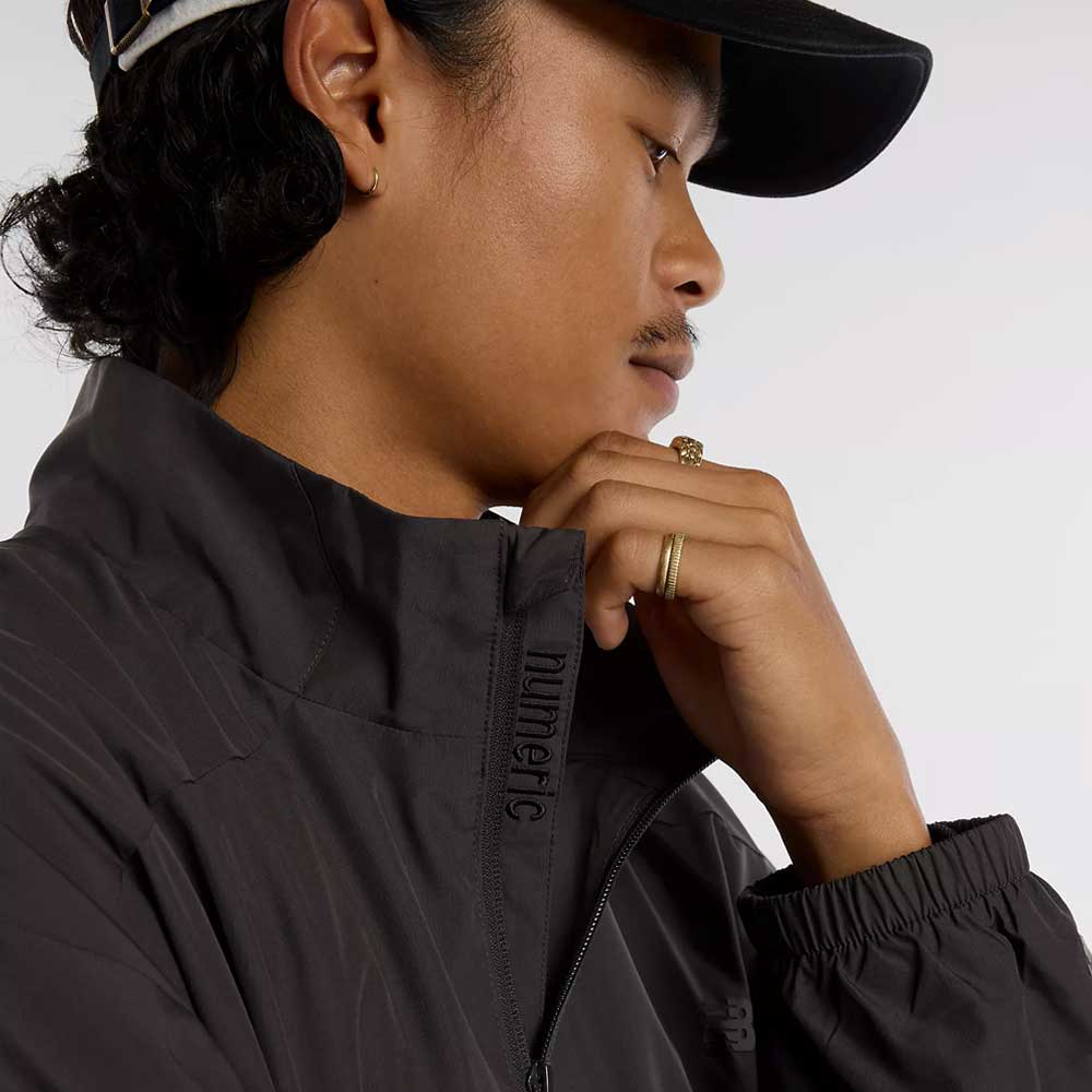 A person in profile wears a black cap and the New Balance Numeric Jacket MJ61W4XCABR in ripstop fabric, with "numeric" near the zipper. They rest their hand on their chin, showcasing gold rings and an earring.