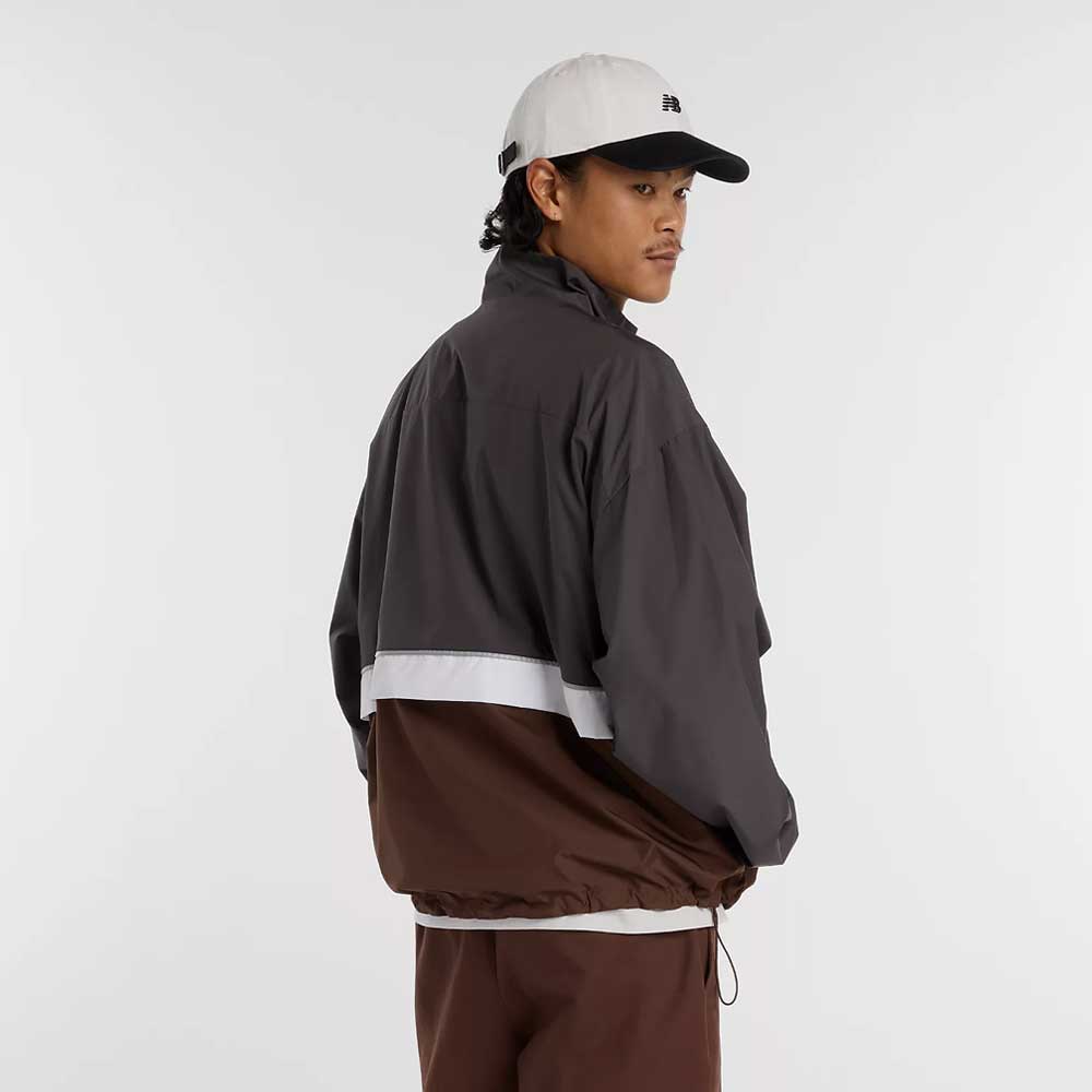 A man wearing a white baseball cap, brown pants, and the New Balance Numeric Jacket MJ61W4XCABR with NB DRY technology and a white stripe stands against a plain white background, looking over his shoulder.