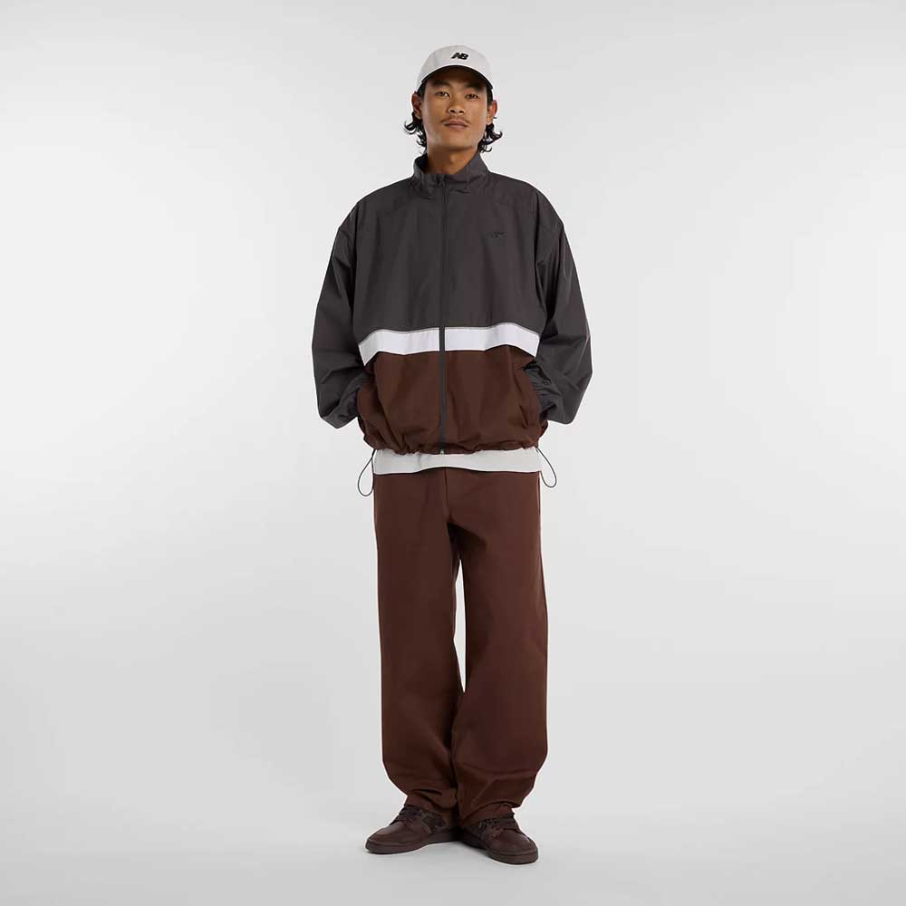 A person faces forward in a light gray cap, wide-legged brown pants, and the New Balance Numeric Jacket MJ61W4XCABR—a color-blocked black, gray, and brown jacket with a white stripe and ripstop fabric. The background is plain and light-colored.