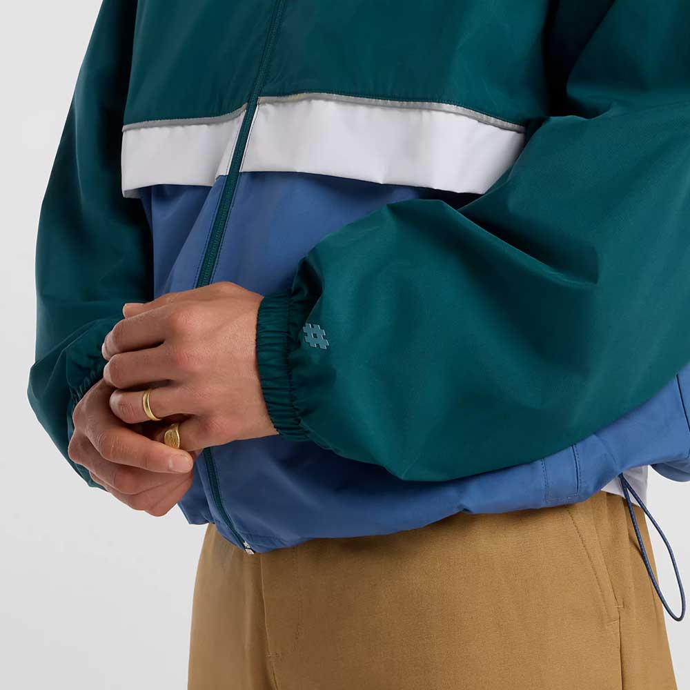 A person wears the New Balance Numeric Jacket MJ61W4XCAAV in teal, blue, and white ripstop fabric with tan pants. Their gold ring-clad hands are clasped, showing only torso and hands against a plain white background.