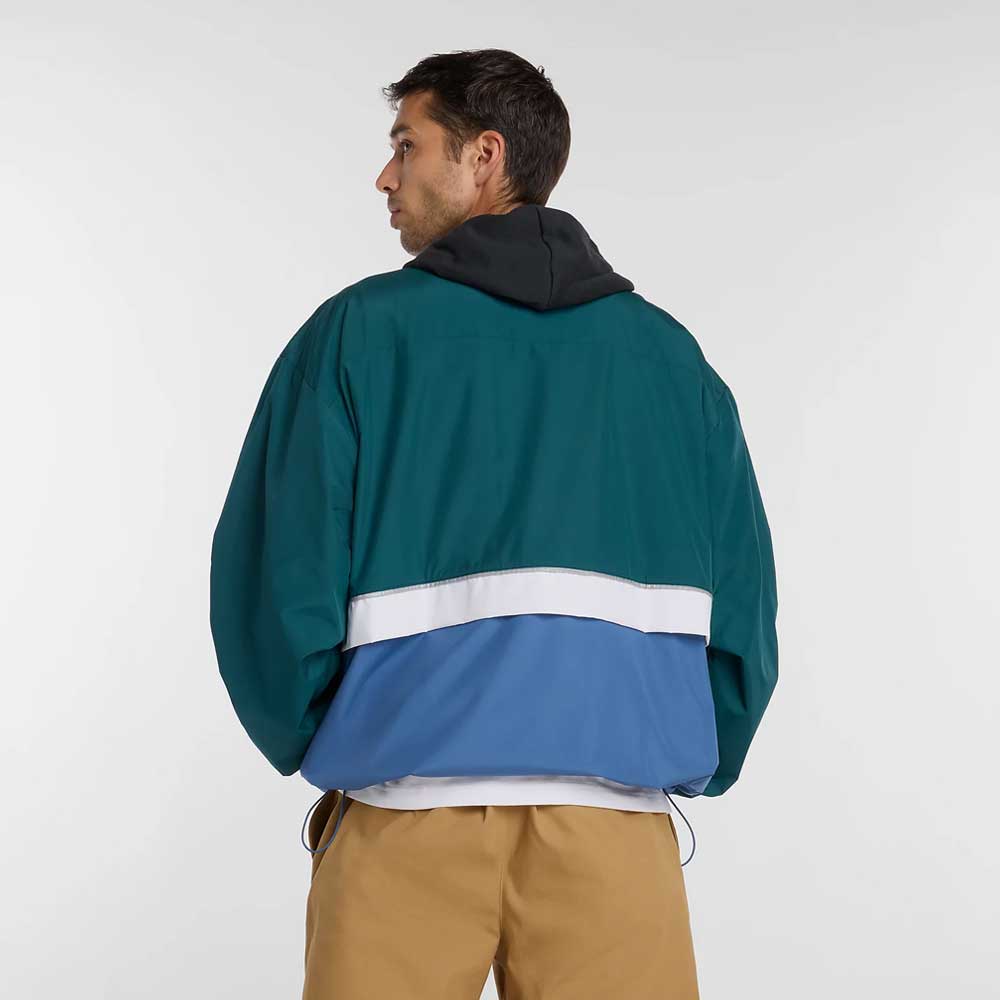 A man faces away wearing the New Balance Numeric Jacket MJ61W4XCAAV in teal, blue, and white color-blocked ripstop fabric with a black hood and tan pants, set against a plain light background.