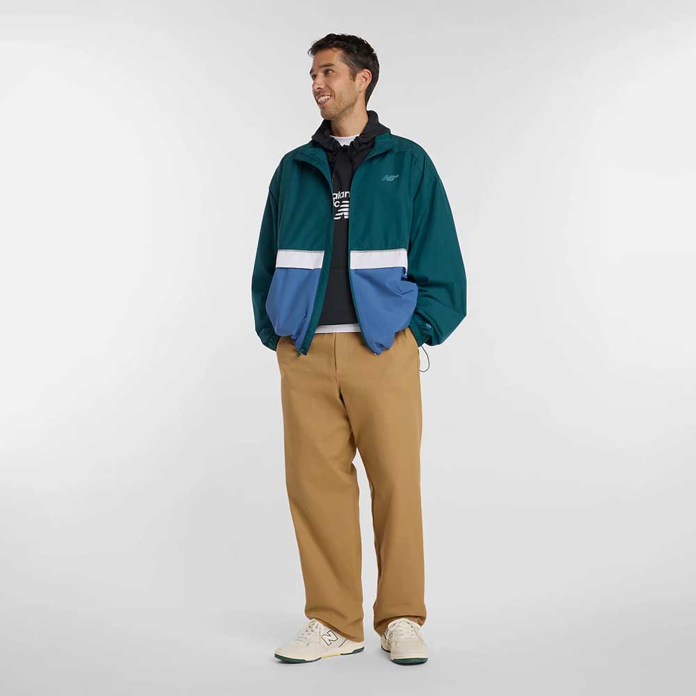 A man stands against a plain background wearing a teal and blue New Balance Numeric Jacket MJ61W4XCAAV made from durable ripstop fabric, a black hoodie with a white logo, tan pants, and white sneakers. He smiles and looks to the side with his hands in his jacket pockets.