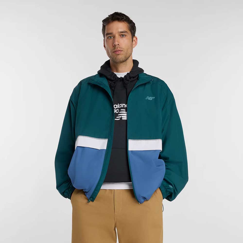 A man with a neutral expression stands against a plain background wearing the New Balance Numeric Jacket MJ61W4XCAAV in teal and blue ripstop fabric over a black hoodie with white lettering, paired with tan pants and his hands in his jacket pockets.