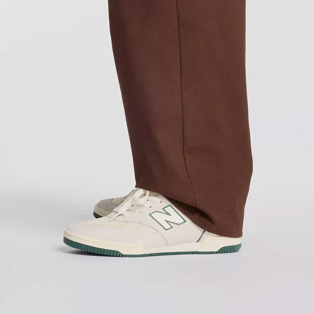 A person wearing New Balance Numeric Pant Pumperknickel MP51953ABJ and white New Balance sneakers with green details stands on a light grey background.