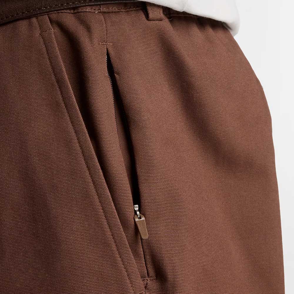 Close-up of New Balance Numeric Pant Pumperknickel MP51953ABJ in brown cotton canvas with a zippered side pocket, textured fabric, and a white shirt tucked in—perfect straight leg pants for a polished yet relaxed look.