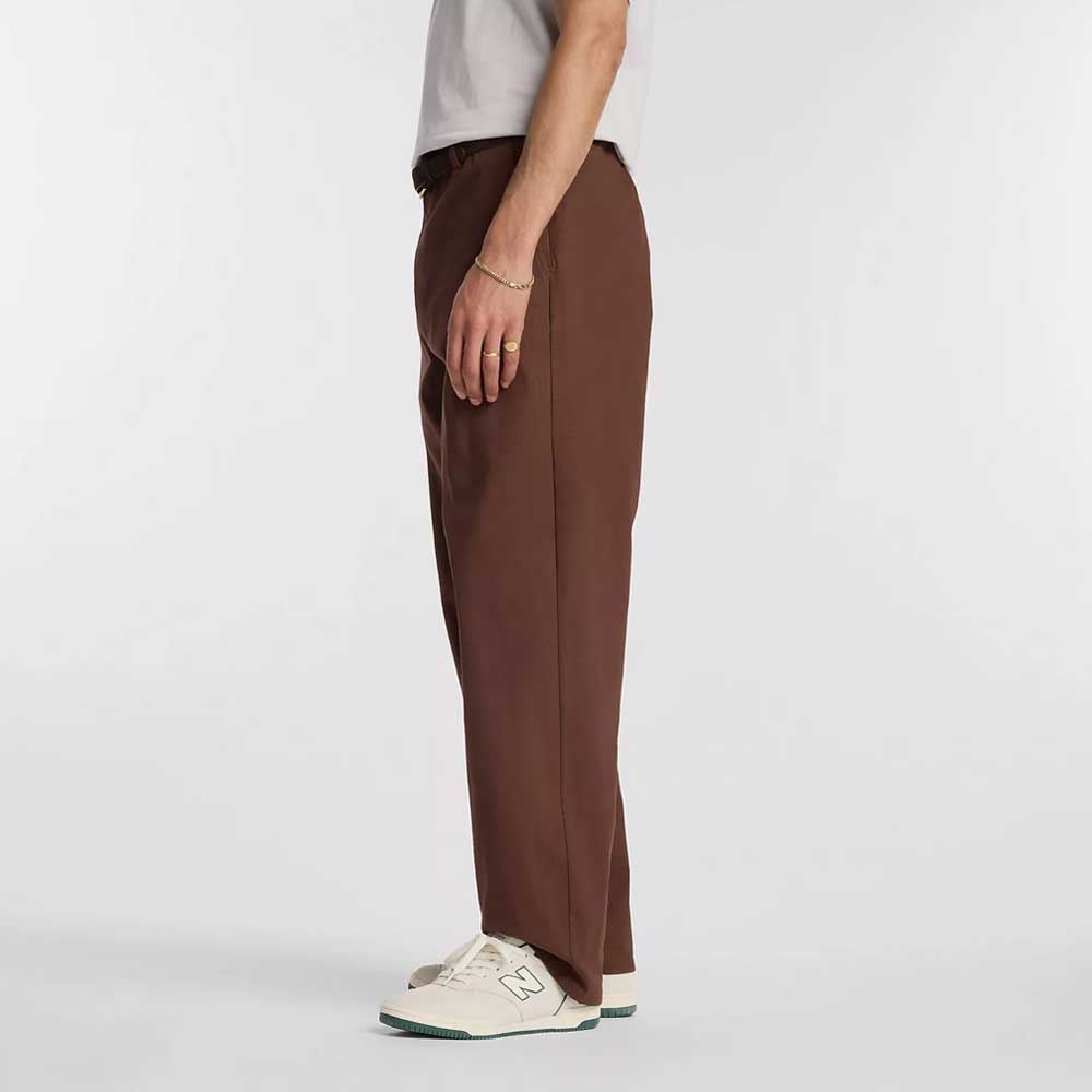 A person stands in profile against a plain white background, wearing the New Balance Numeric Pant Pumperknickel MP51953ABJ, a light t-shirt, a bracelet, and white New Balance sneakers.