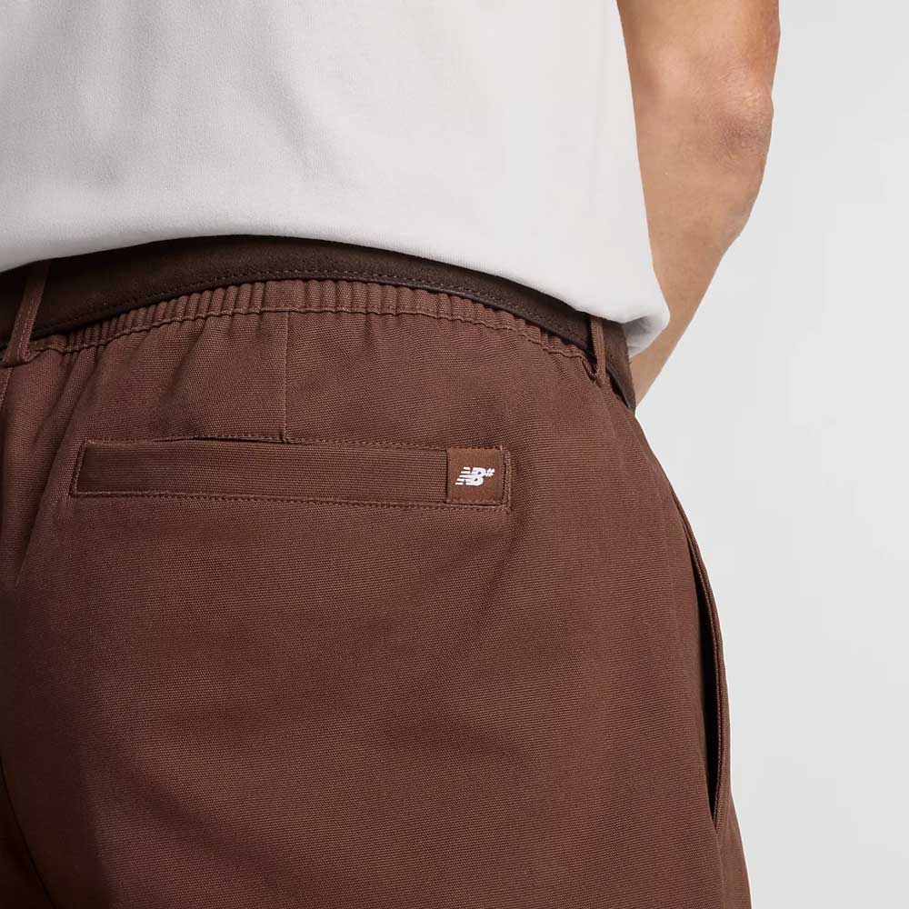 Close-up of a person wearing the New Balance Numeric Pant Pumperknickel MP51953ABJ, featuring brown cotton canvas, an elastic waistband, and a small logo tag above the back pocket, paired with a tucked-in white shirt.