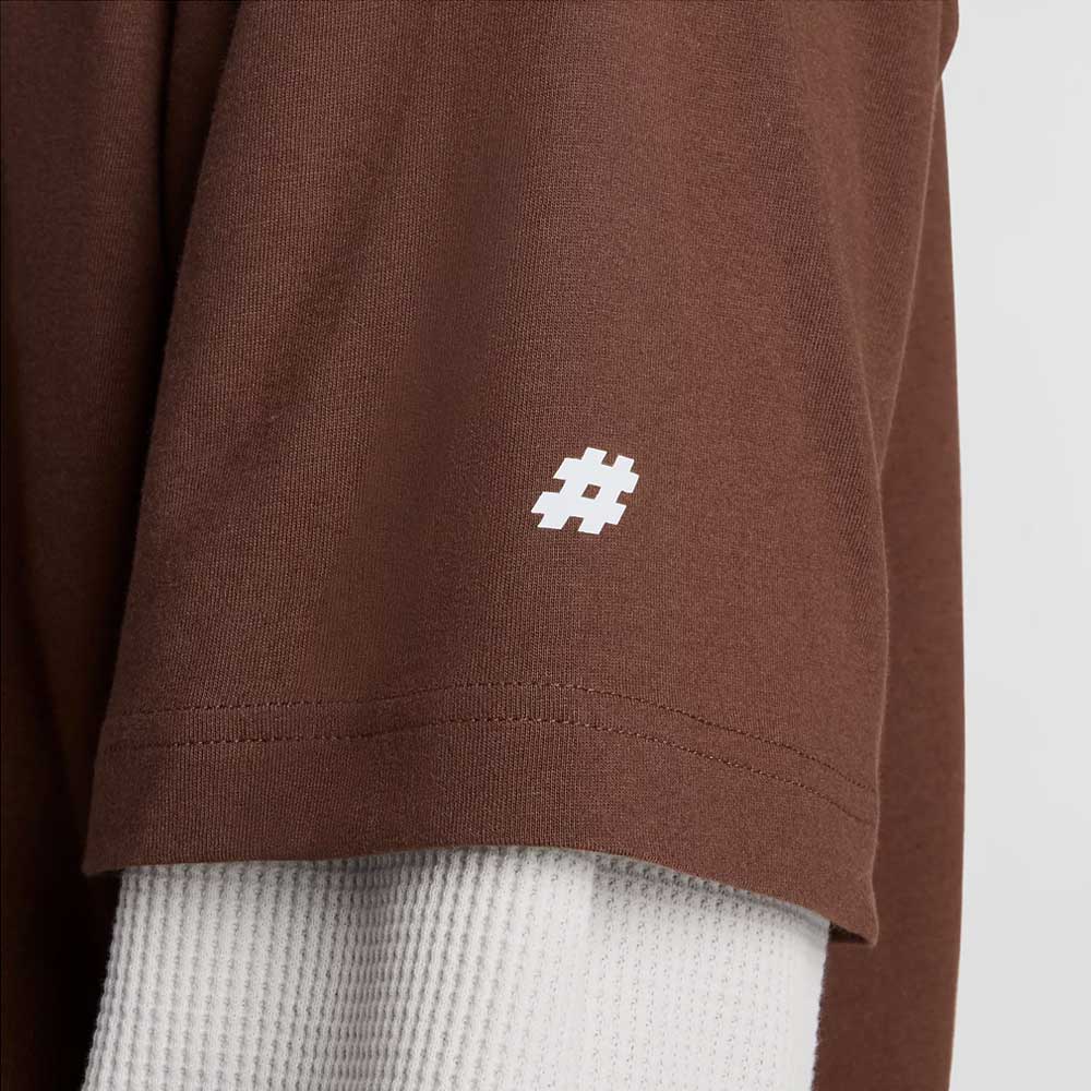 A close-up of the New Balance Numeric Small Logo Tee Pumperknickel MT53706ABJ, a brown cotton crewneck in a relaxed fit with a small white hashtag logo by the sleeve hem, layered over an off-white ribbed long-sleeve shirt.