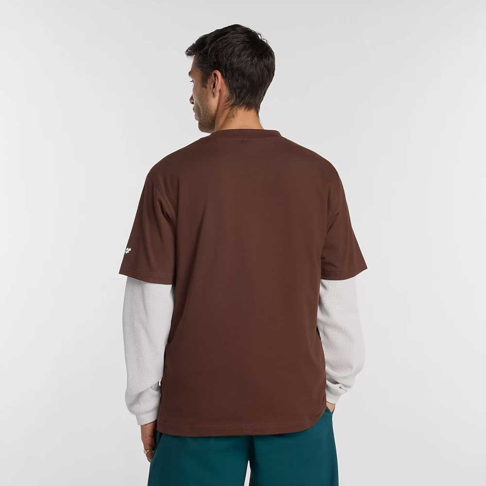 A man faces away from the camera, wearing the New Balance Numeric Small Logo Tee Pumperknickel MT53706ABJ over a white long-sleeve shirt and teal pants with Numeric sneakers, set against a plain light gray background.