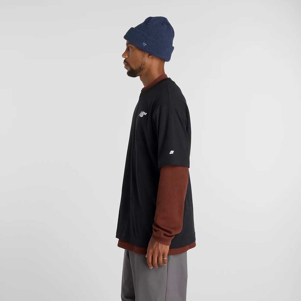 A man in relaxed fit gray pants, a blue knit beanie, and a New Balance Numeric Small Logo Black Tee MT53706BK layered over a brown long-sleeve shirt stands in profile against a plain light background.