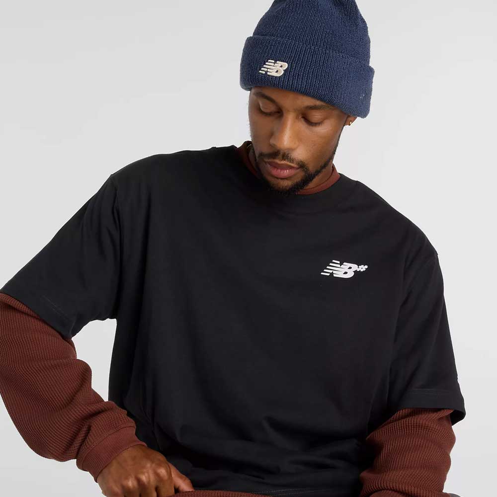 A man in a New Balance Numeric Small Logo Black Tee MT53706BK over a long-sleeve brown shirt and a blue New Balance beanie looks down with a neutral expression against a plain background.