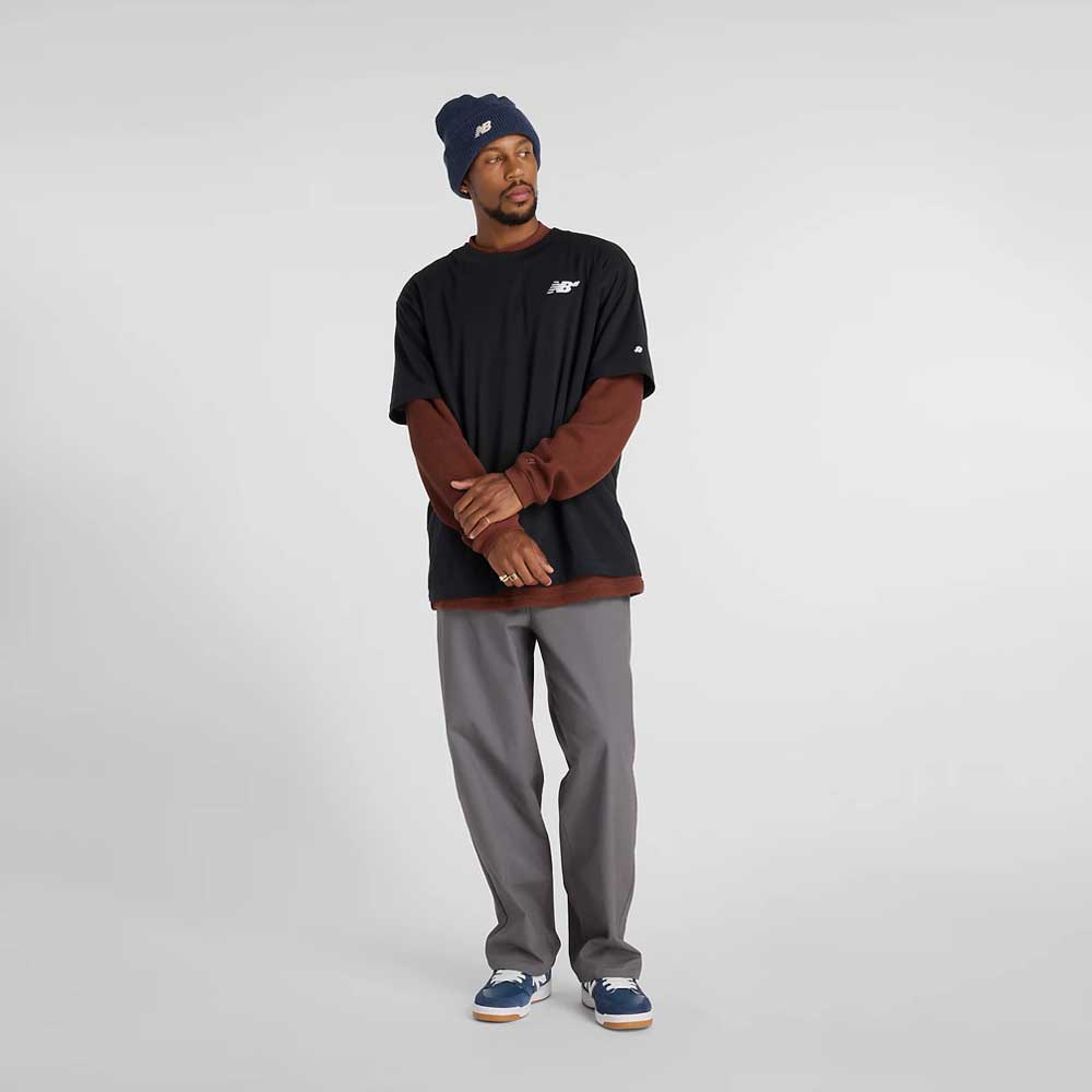 A man wears a navy beanie, New Balance Numeric Small Logo Black Tee MT53706BK over a brown long-sleeve shirt, relaxed fit gray pants, and blue New Balance Numeric sneakers against a plain light gray background.