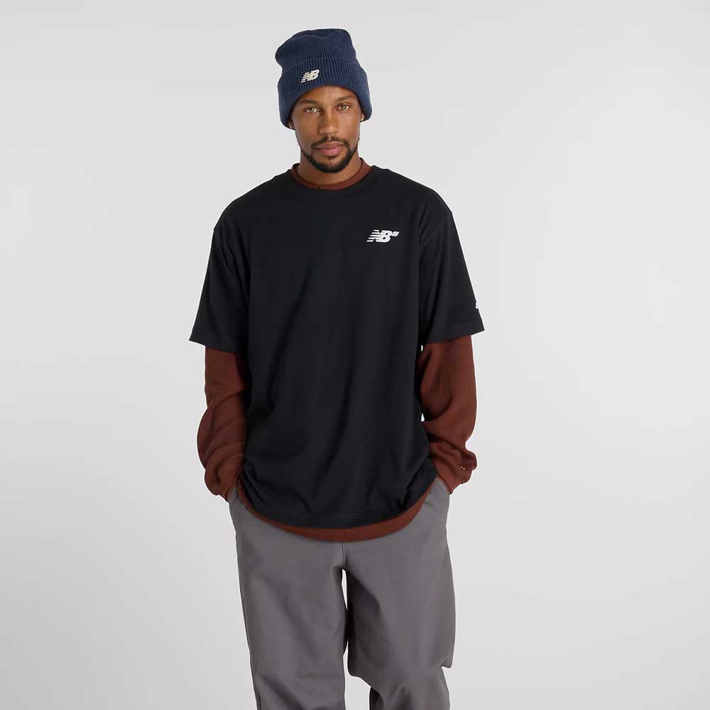A man in a navy beanie, gray pants, and a relaxed fit New Balance Numeric Small Logo Black Tee MT53706BK over a brown long-sleeve shirt stands with hands in pockets against a plain light background.