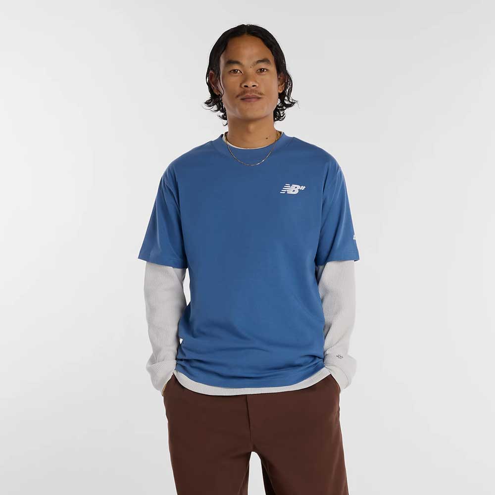 A person with wavy dark hair wears the New Balance Numeric Small Logo Magic Blue Tee MT53706AA4 over a long-sleeved gray shirt and brown pants, standing with hands in pockets against a plain white background.