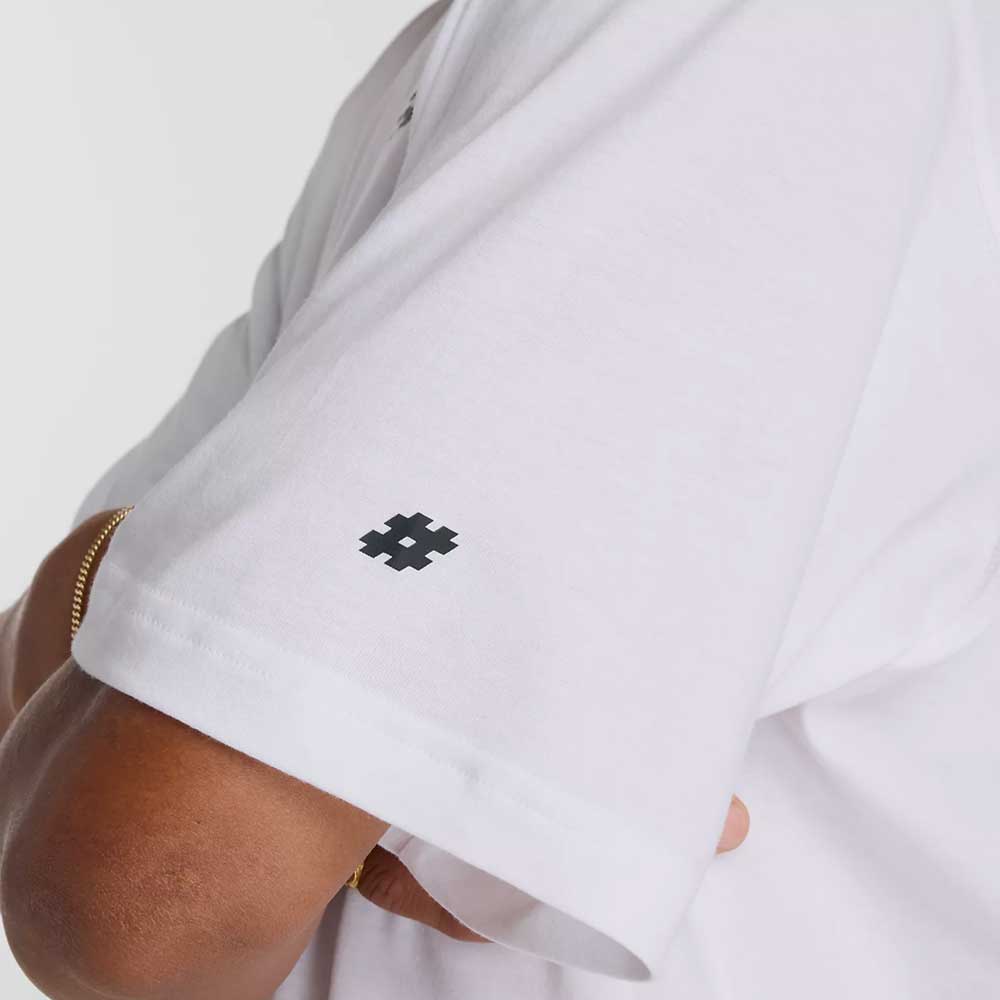 A close-up of a person’s crossed arms in the New Balance Numeric Small Logo White Tee MT53706WT, a relaxed fit cotton crewneck featuring a small black pixel-style hashtag symbol on the sleeve.
