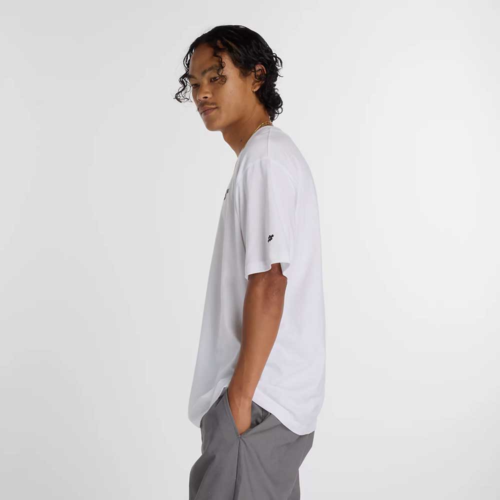 Young person with curly hair stands in profile, hands in pockets, wearing the New Balance Numeric Small Logo White Tee MT53706WT and gray pants, paired with New Balance Numeric sneakers against a plain light background.