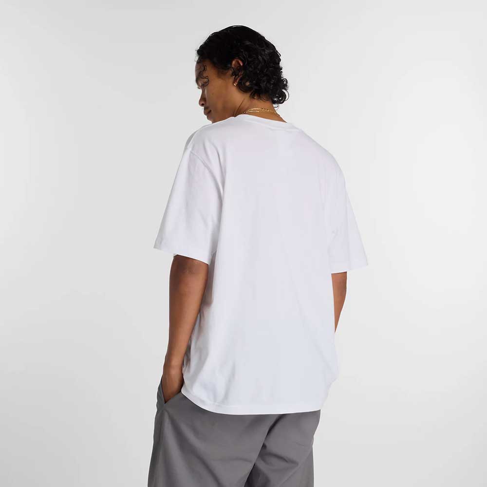 A person with short, curly hair faces away, wearing the New Balance Numeric Small Logo White Tee MT53706WT and gray pants, hands in their pockets, set against a plain white background.