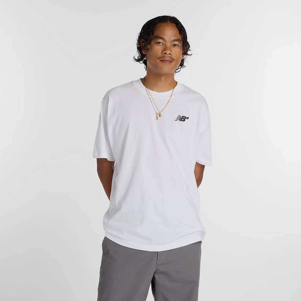 A person with short curly hair wears the New Balance Numeric Small Logo White Tee MT53706WT in a relaxed fit, paired with gray pants and gold necklaces, standing with hands behind their back against a plain white background.