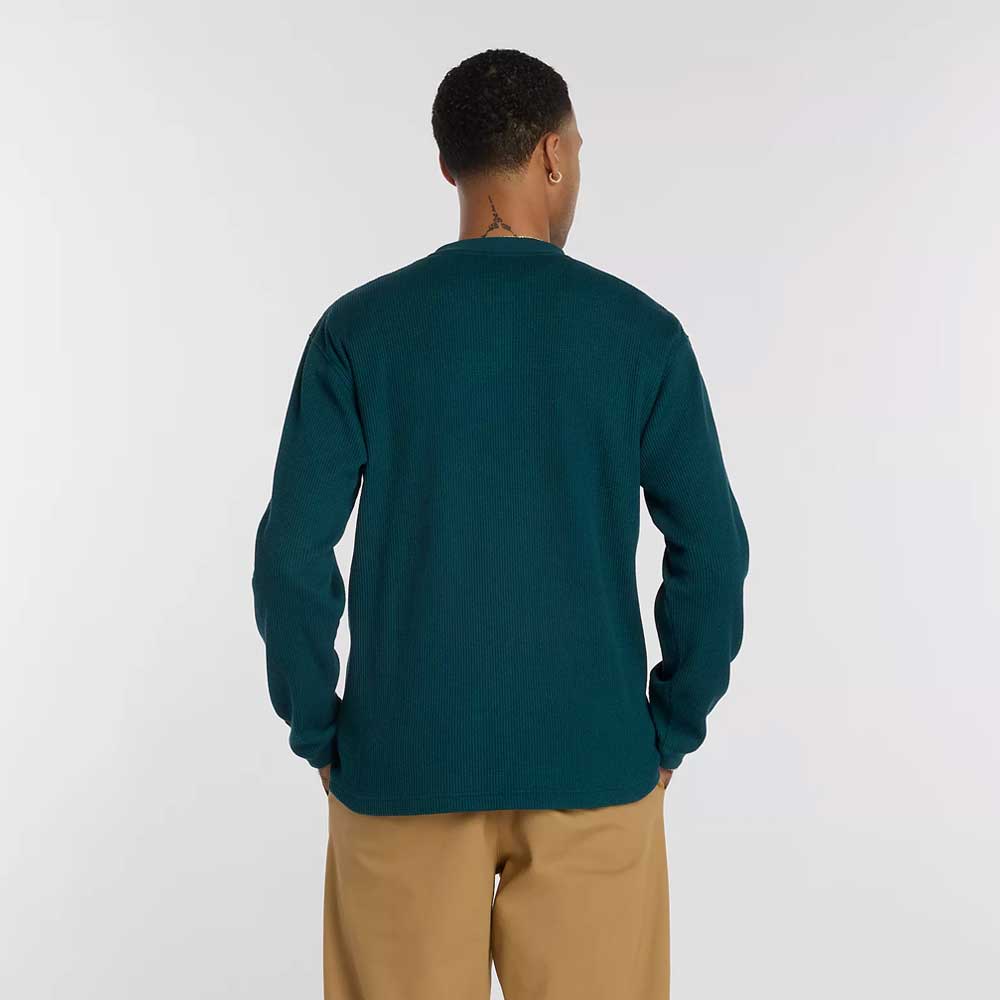 A person faces away from the camera, wearing a New Balance Numeric Waffle Knit Tee MT51958AAV in dark teal and tan pants against a plain, light background.